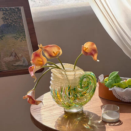 Modern Coastal Chic hand-blown art glass nautilus shell vase with soft-hued flowers on wooden table, featuring spiral patterns in green and yellow tones, natural light from window.