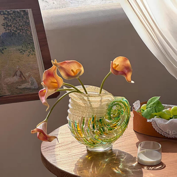 Modern Coastal Chic hand-blown art glass nautilus shell vase with soft-hued flowers on wooden table, featuring spiral patterns in green and yellow tones, natural light from window.