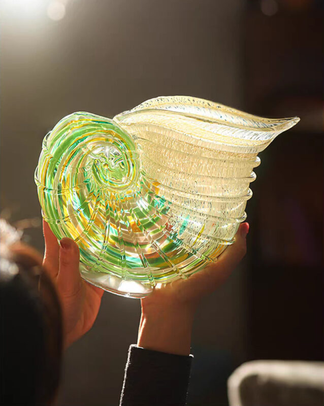 Hands holding Coastal Chic art glass conch shell vase with green-yellow gradient spiral patterns, glowing under dramatic top-left lighting against dark backdrop.