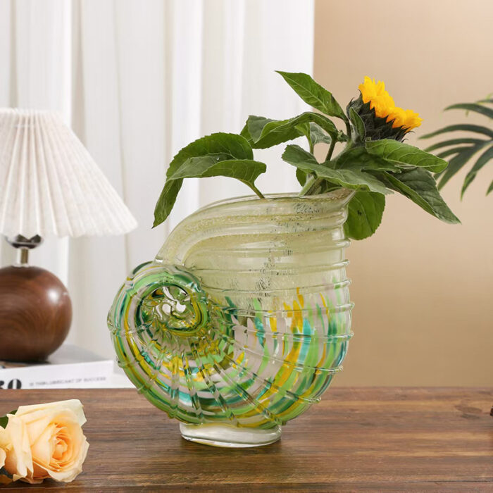 Hand-blown conch shell glass vase with green-yellow stripes, holding yellow flowers and pink roses on wooden table, styled with pleated lamp and sheer curtains in natural light.