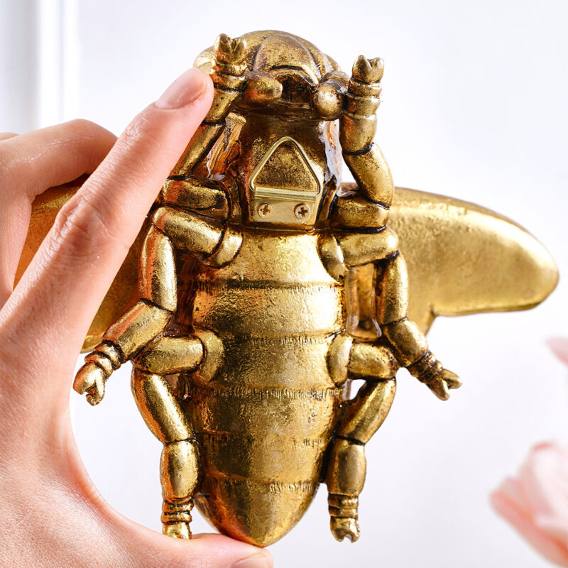Golden resin bumblebee wall sculpture with steampunk goggles and jointed mechanical arms, held by hand against white background