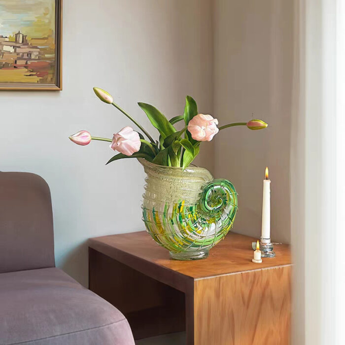 ​：Decorative spiral shell glass vase in soft green tones with pink tulips and foliage on wooden side table, white candles on right, abstract landscape painting on light wall, grey sofa in cozy interior.