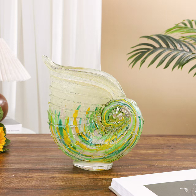 Nautilus shell-shaped art glass centerpiece vase in pale yellow with green-yellow spirals on wooden table, styled with white curtains and greenery in natural-lit coastal chic interior.