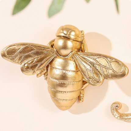 Golden resin bee wall art with 3D relief details on light beige background, featuring decorative scroll element