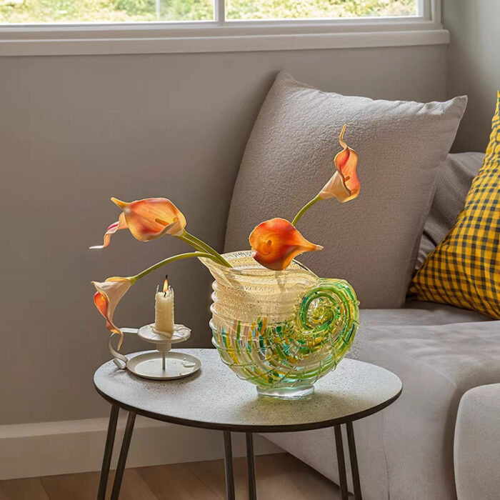 Hand-blown spiral art glass vase with green-yellow gradients, holding orange calla lilies and candle on round wooden table, staged against light grey sofa and window greenery.