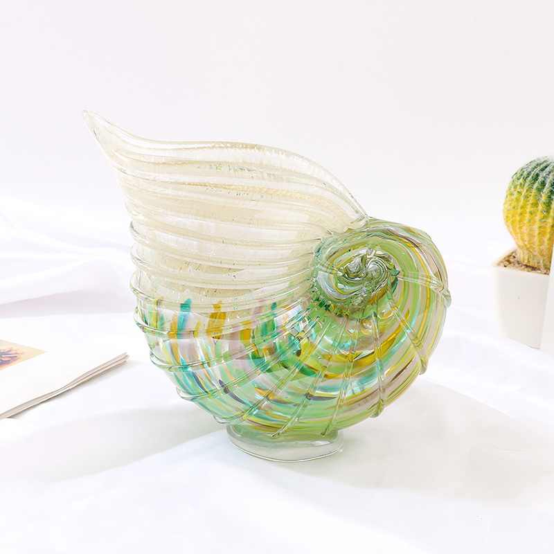 Hand-blown seashell-shaped art glass vase with transparent upper section and green-yellow-blue gradients, staged with succulent plant and open book on white backdrop.