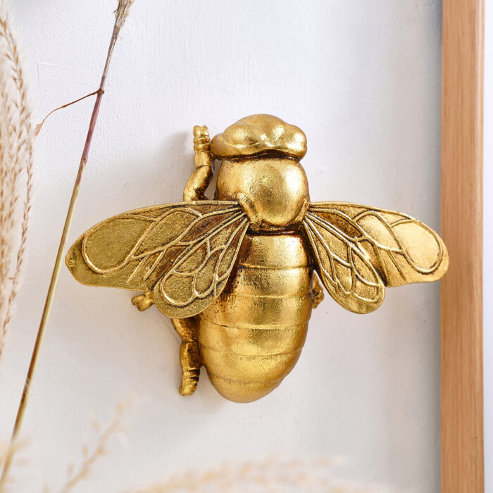 Golden honey bee resin wall sculpture with anatomical wings and segmented abdomen, framed by natural wood border and dried grasses on white wall