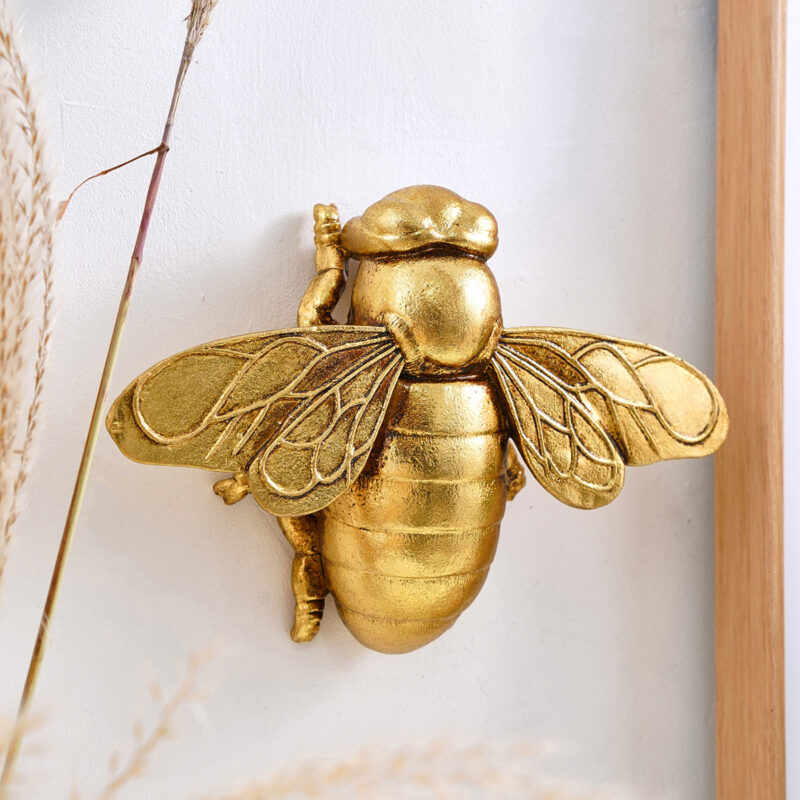 Golden honey bee resin wall sculpture with anatomical wings and segmented abdomen, framed by natural wood border and dried grasses on white wall