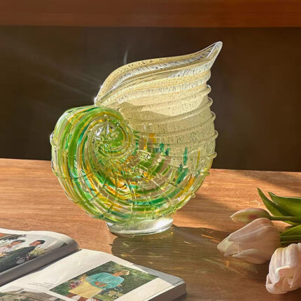 Hand-blown nautilus shell art glass vase with translucent amber-toned exterior and green-yellow spiral gradients, staged with pale pink tulips & books on wooden table under warm side lighting.
