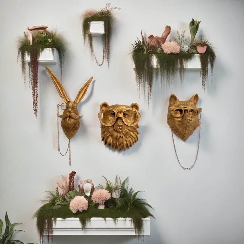 Three gold animal head wall decor (bear/fox/rabbit) with glasses & chains: monocle-wearing mustachioed rabbit, bear & fox in oversized frames, displayed on light gray wall with white floating shelves holding trailing plants & pink blooms | Whimsical nature-inspired wall art
